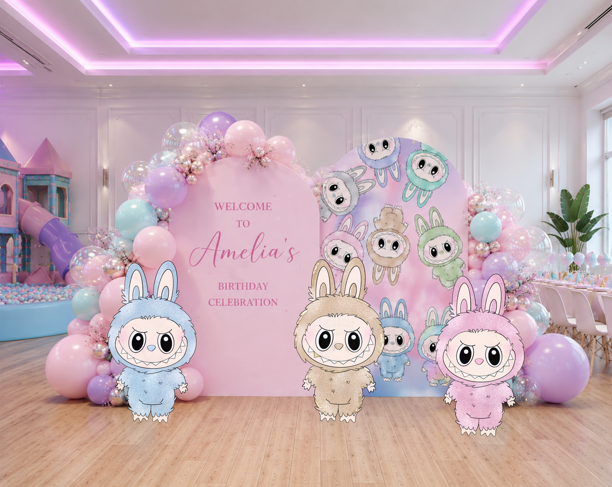 Indoor pastel monster party backdrop with soft pink and lavender panels, balloon garland and cute monster cutouts for a personalized birthday.