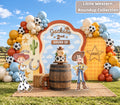 Personalized western toy birthday backdrop with cowboy-inspired character cutouts, hay bales, rustic barrels and warm orange blue balloon decor.