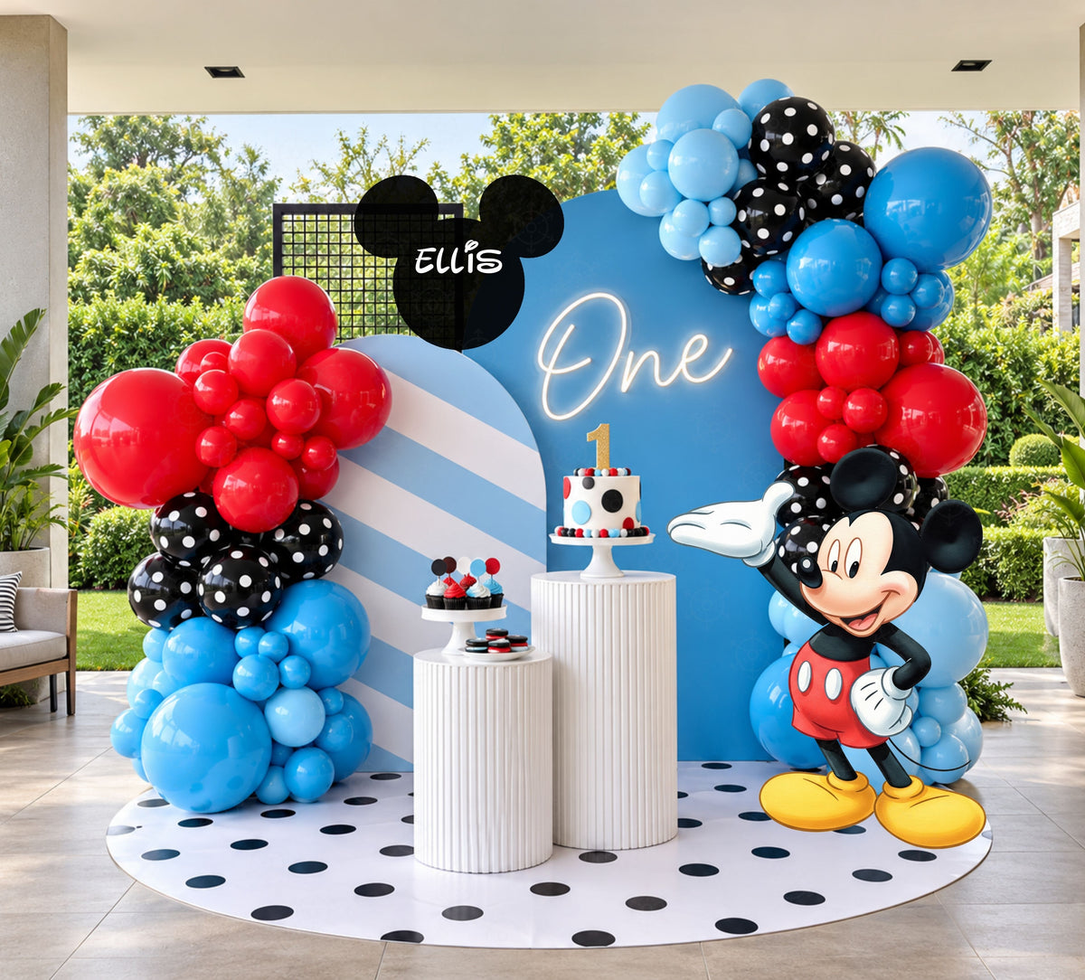 Classic mouse first birthday backdrop with blue arched panel, red blue and black balloon garland, personalized name sign and matching character cutout.